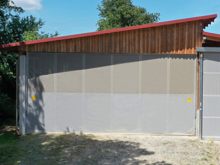 Manuelles Schiebetor als Aus- und Einfahrt zu einer landwirtschaftlichen Maschinenhalle. Materialien: Windschutznetz und Plane.